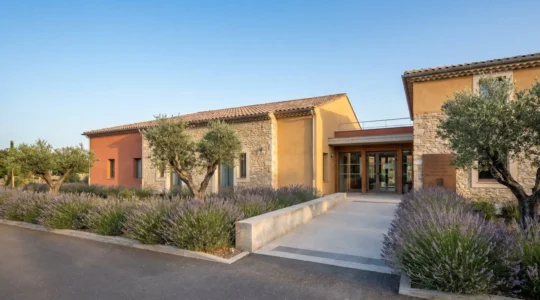 Façade d'un établissement EHPAD de style provençal avec entrée accessible, entouré de végétation méditerranéenne sous ciel bleu